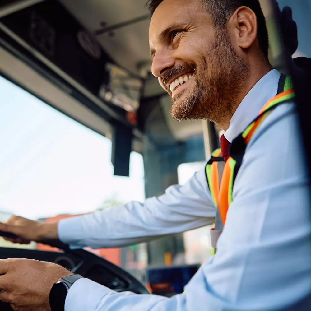 Een chauffeur in een nette shirt, stropdas en oranje veiligheids hesje lacht terwijl hij een vrachtwagen bestuurt.
